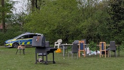 Auf einer Wiese stehen verteilt verschiedene Stühle, ein Esstisch und ein Grill. Im Hintergrund liegt Müll auf dem Boden. Ein Polizeiauto steht in der linken Bildecke.